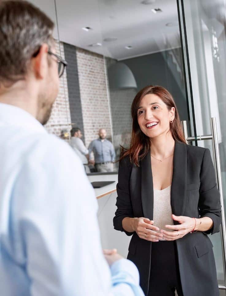 Frau mit rot-braunen Haaren in Business Outfit im Gespräch mit einem Kollegen.